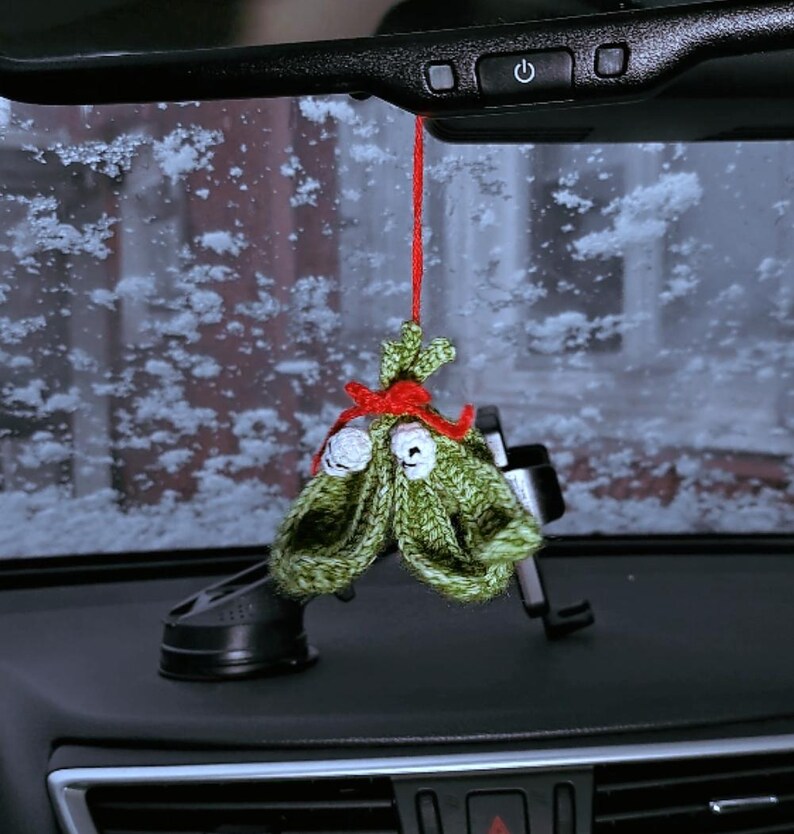 Pu&ograve; includere: Un ornamento di Natale a forma di albero di Natale in crochet verde con accenti rossi e bianchi appeso a uno specchietto retrovisore in un'auto.