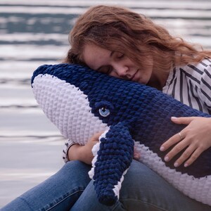 K&ouml;nnte beinhalten: Ein gro&szlig;es, handgeh&auml;keltes Wal-Kissen in Marineblau und Wei&szlig;. Das Pl&uuml;schtier hat ein detailliertes Auge und Flossen. Der Wal wird von einer Person gehalten, die eine blaue Jeans und ein gestreiftes Hemd tr&auml;gt. Der Hintergrund ist eine verschwommene Ansicht von Wasser.