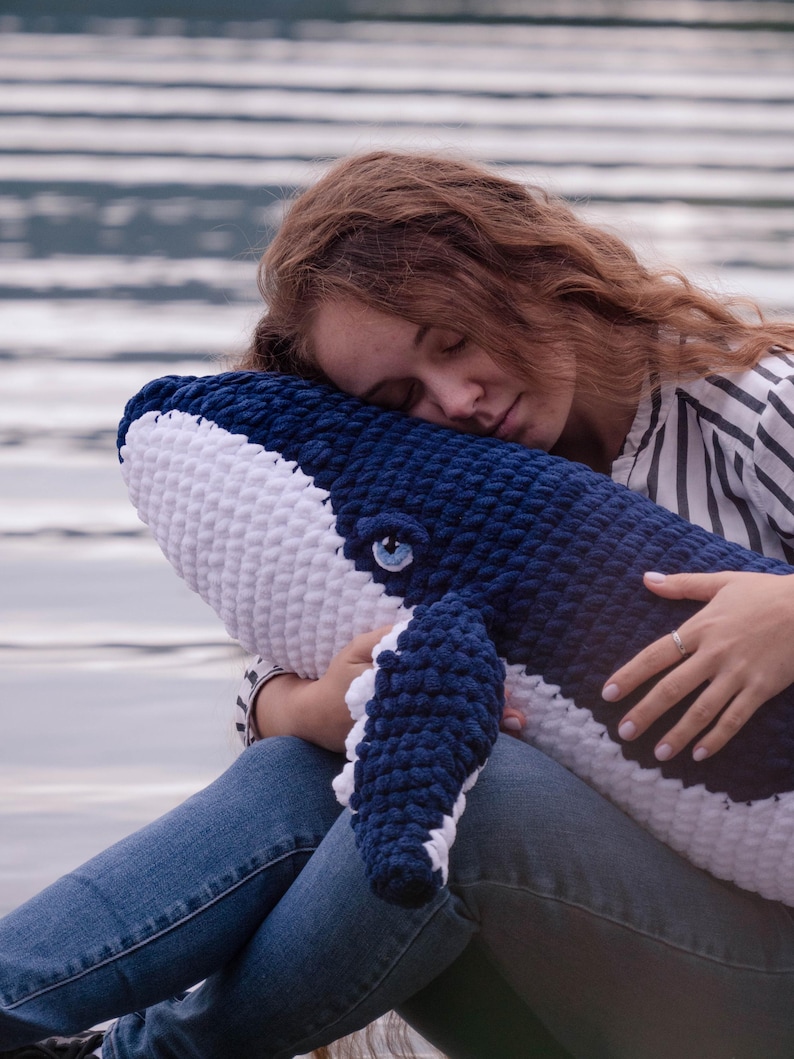 K&ouml;nnte beinhalten: Ein gro&szlig;es, handgeh&auml;keltes Wal-Kissen in Marineblau und Wei&szlig;. Das Pl&uuml;schtier hat ein detailliertes Auge und Flossen. Der Wal wird von einer Person gehalten, die eine blaue Jeans und ein gestreiftes Hemd tr&auml;gt. Der Hintergrund ist eine verschwommene Ansicht von Wasser.