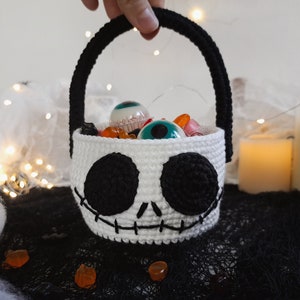 K&ouml;nnte beinhalten: Ein wei&szlig;er geh&auml;kelter Halloween-Trick-or-Treat-Korb in Form von Jack Skellingtons Gesicht. Der Korb hat schwarze Augen, einen gestickten Mund und einen schwarzen Griff. Er ist gef&uuml;llt mit S&uuml;&szlig;igkeiten und Spielzeugaugen.