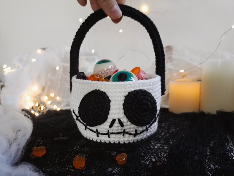 K&ouml;nnte beinhalten: Ein wei&szlig;er geh&auml;kelter Halloween-Trick-or-Treat-Korb in Form von Jack Skellingtons Gesicht. Der Korb hat schwarze Augen, einen gestickten Mund und einen schwarzen Griff. Er ist gef&uuml;llt mit S&uuml;&szlig;igkeiten und Spielzeugaugen.