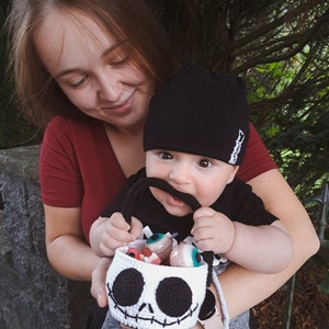 K&ouml;nnte beinhalten: Ein Baby mit einer schwarzen M&uuml;tze und einem schwarz-wei&szlig;en Shirt h&auml;lt eine geh&auml;kelte Jack Skellington-Trick-or-Treat-Tasche, die mit S&uuml;&szlig;igkeitenaugen gef&uuml;llt ist. Das Baby wird von einer Frau in einem roten Shirt gehalten.