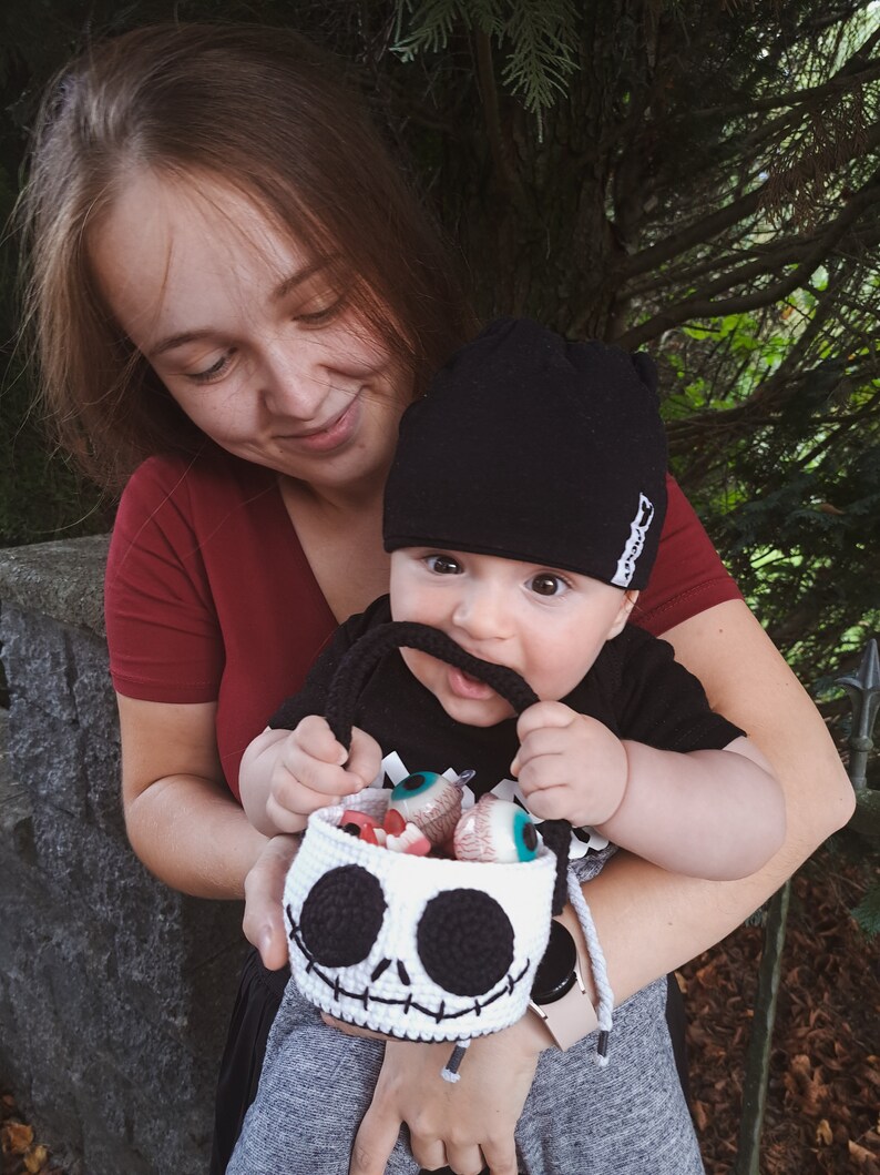 K&ouml;nnte beinhalten: Ein Baby mit einer schwarzen M&uuml;tze und einem schwarz-wei&szlig;en Shirt h&auml;lt eine geh&auml;kelte Jack Skellington-Trick-or-Treat-Tasche, die mit S&uuml;&szlig;igkeitenaugen gef&uuml;llt ist. Das Baby wird von einer Frau in einem roten Shirt gehalten.