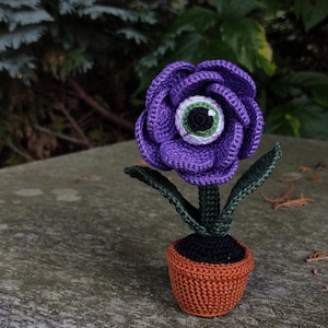 Puede incluir: Una flor de color morado tejida a crochet con un ojo verde en el centro, sentada en una maceta marr&oacute;n con tierra negra. La flor tiene hojas verdes.