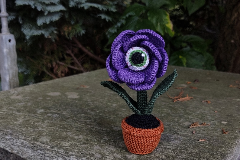Puede incluir: Una flor de color morado tejida a crochet con un ojo verde en el centro, sentada en una maceta marr&oacute;n con tierra negra. La flor tiene hojas verdes.