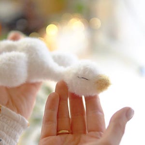 May include: A soft, white plush goose toy with a yellow beak and a closed eye. The toy is held in a person's hands, with a gold ring visible on one finger. The background is blurred, suggesting a soft focus.