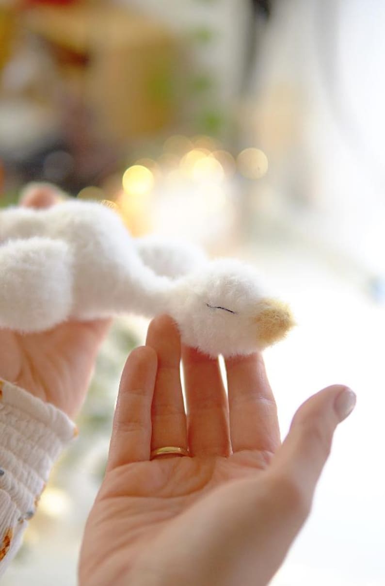 May include: A soft, white plush goose toy with a yellow beak and a closed eye. The toy is held in a person's hands, with a gold ring visible on one finger. The background is blurred, suggesting a soft focus.