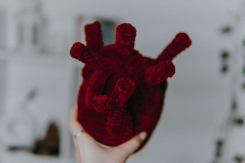 May include: A plush red heart shaped toy made of yarn. The heart has a textured surface and is held in a hand.