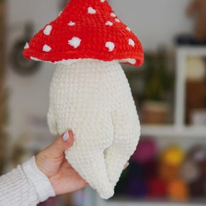 以下が含まれることがあります： 手作りのキノコのおもちゃ。赤い帽子には白い水玉模様が施され、茎はオフホワイトです。おもちゃは柔らかいぬいぐるみ素材で作られており、漫画のキャラクターのようにデザインされています。
