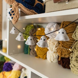 Puede incluir: Un estante exhibe una variedad de suministros para manualidades. Una tortuga de ganchillo se encuentra en la parte superior. Una guirnalda de peque&ntilde;as decoraciones de fantasmas blancos cuelga frente a bolas de hilo coloridas. El hilo es en tonos verdes, amarillos, blancos y marrones.