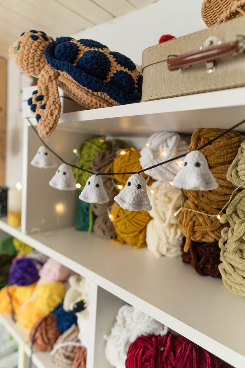 Puede incluir: Un estante exhibe una variedad de suministros para manualidades. Una tortuga de ganchillo se encuentra en la parte superior. Una guirnalda de peque&ntilde;as decoraciones de fantasmas blancos cuelga frente a bolas de hilo coloridas. El hilo es en tonos verdes, amarillos, blancos y marrones.