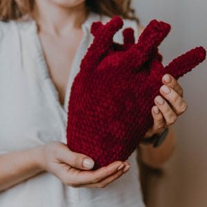 May include: A woman holds a crocheted heart-shaped plush toy in her hands. The toy is a deep red color and has a textured surface.
