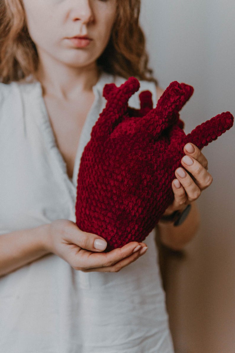 May include: A woman holds a crocheted heart-shaped plush toy in her hands. The toy is a deep red color and has a textured surface.