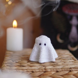K&ouml;nnte beinhalten: Ein wei&szlig;er geh&auml;kelter Geist mit schwarzen Augen sitzt auf einer geflochtenen Weidenoberfl&auml;che. Der Geist ist eine Halloween-Dekoration.