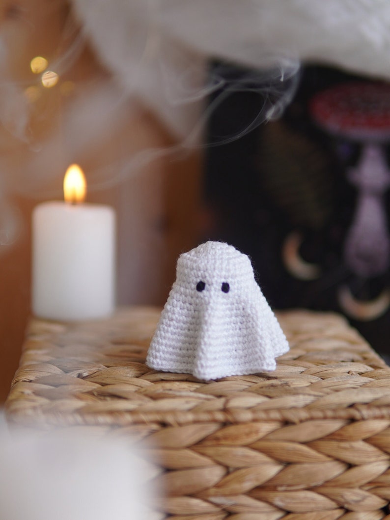 K&ouml;nnte beinhalten: Ein wei&szlig;er geh&auml;kelter Geist mit schwarzen Augen sitzt auf einer geflochtenen Weidenoberfl&auml;che. Der Geist ist eine Halloween-Dekoration.