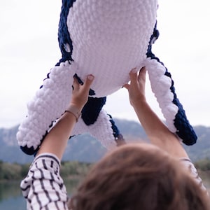 K&ouml;nnte beinhalten: Ein gro&szlig;es, kuscheliges Walspielzeug in Wei&szlig; und Marineblau. Der Wal wird vor dem Hintergrund eines Sees und von Bergen gehalten. Das Spielzeug besteht aus weichem, strukturiertem Material und die Farben sind lebendig.