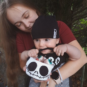 K&ouml;nnte beinhalten: Eine Frau h&auml;lt ein Baby, das eine schwarze M&uuml;tze mit dem Wort "Mickey" tr&auml;gt. Das Baby h&auml;lt einen geh&auml;kelten Sch&auml;delkorb, der mit bunten Aug&auml;pfeln gef&uuml;llt ist. Das Baby tr&auml;gt ein schwarz-graues Shirt mit der Nummer "28" darauf.