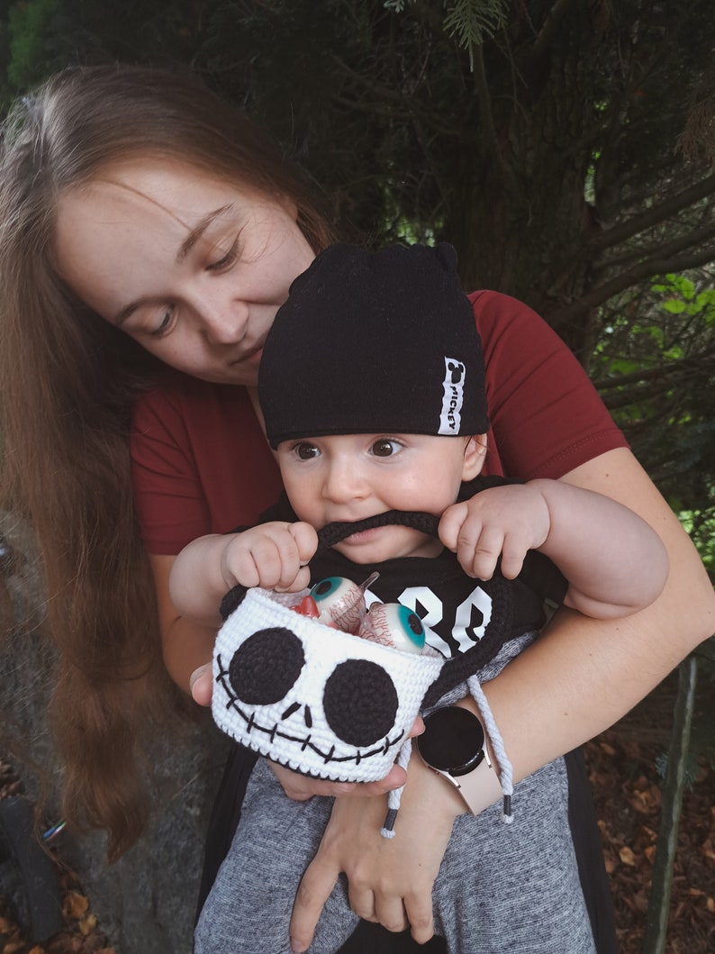 K&ouml;nnte beinhalten: Eine Frau h&auml;lt ein Baby, das eine schwarze M&uuml;tze mit dem Wort "Mickey" tr&auml;gt. Das Baby h&auml;lt einen geh&auml;kelten Sch&auml;delkorb, der mit bunten Aug&auml;pfeln gef&uuml;llt ist. Das Baby tr&auml;gt ein schwarz-graues Shirt mit der Nummer "28" darauf.