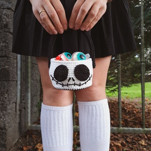 K&ouml;nnte beinhalten: Eine wei&szlig;e geh&auml;kelte Trick-or-Treat-Tasche in Form von Jack Skellingtons Gesicht aus Nightmare Before Christmas. Die Tasche ist mit bunten Gummiaugen gef&uuml;llt.