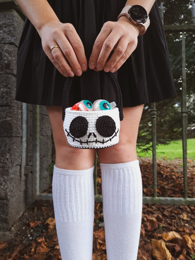 K&ouml;nnte beinhalten: Eine wei&szlig;e geh&auml;kelte Trick-or-Treat-Tasche in Form von Jack Skellingtons Gesicht aus Nightmare Before Christmas. Die Tasche ist mit bunten Gummiaugen gef&uuml;llt.