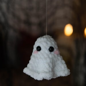 Puede incluir: Un fantasma blanco de ganchillo hecho a mano con ojos de bot&oacute;n negros y mejillas rosadas. El fantasma est&aacute; suspendido por un hilo fino, con un fondo borroso. Esta decoraci&oacute;n caprichosa es perfecta para Halloween o para todo el a&ntilde;o.