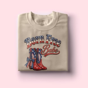May include: A beige sweatshirt featuring a graphic with the text "Born Free Babe". The graphic includes a pair of red cowboy boots with blue star accents and a blue bow. The text is in red, white, and blue.