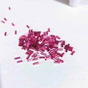 May include: A pile of pink glass beads in the shape of small, thin cylinders. The beads are scattered on a white surface.