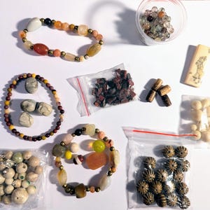 Puede incluir: Surtido de cuentas y suministros para la elaboración de joyas en varios colores y materiales, incluyendo blanco, marrón, beige, rojo, naranja, amarillo y verde. Los suministros están en pequeñas bolsas y contenedores de plástico.