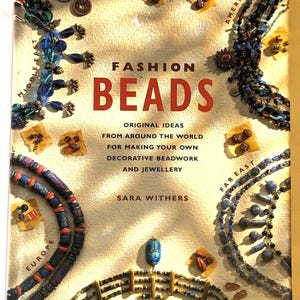 Może przedstawiać: Książka zatytułowana "Fashion Beads" z kolorową okładką przedstawiającą różne koraliki i biżuterię z całego świata. Książka dotyczy tworzenia własnych dekoracyjnych prac z koralików i biżuterii.