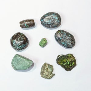 May include: An assortment of polished stones in various shapes and colors. The stones include shades of green, brown, and black, with turquoise accents. The arrangement is circular, showcasing the different textures and hues of the natural gemstones.