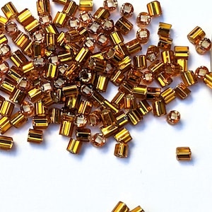 May include: A pile of small, gold-colored glass beads. The beads are cylindrical in shape and have a shiny, metallic finish.
