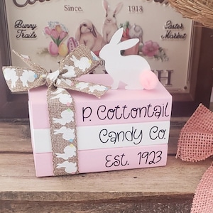 May include: A stack of three pink painted wooden blocks with a white bunny cutout on top. The blocks are decorated with a burlap ribbon with white bunny silhouettes and colorful polka dots. The blocks are labeled "P. Cottontail Candy Co. Est. 1923".
