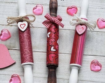 May include: Three wooden rolling pins decorated for Valentine's Day. The rolling pins are wrapped in red and white patterned paper with hearts. One rolling pin has a red and white gingham bow, one has a burlap bow with a heart, and one has a white bow with a heart.