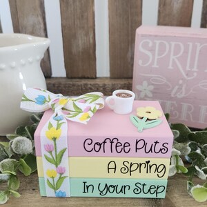May include: A decorative stack of pastel-colored blocks with the text "Coffee Puts A Spring In your Step." A white bow with floral accents and a miniature coffee cup and flower embellish the top. The theme is spring and coffee.