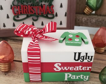 May include: A wooden box with a red and white striped bow, a green sweater with white buttons, and the words "Ugly Sweater Party" written on the front. The box is sitting on a wooden surface.