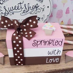 May include: A stack of three wooden boxes with a pink, white, and brown color scheme. The top box has a small cupcake decoration and the text "Sprinkled". The middle box has the text "with" and the bottom box has the text "Love". A brown ribbon with white polka dots is tied around the boxes.