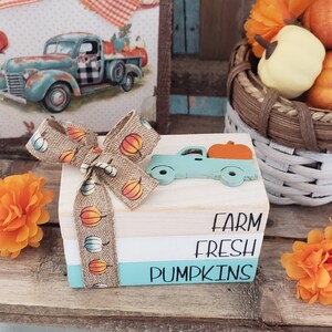 May include: A set of three wooden blocks stacked on top of each other. The top block is white with the word "FARM" written in black. The middle block is white with the word "FRESH" written in black. The bottom block is light blue with the word "PUMPKINS" written in black. A small blue truck with an orange pumpkin in the bed is attached to the top block. A brown burlap ribbon with a pumpkin print is tied around the blocks.