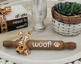 May include: A white tiered tray with a wooden rolling pin that says "woof!" with a paw print. The tray also has a stack of white blocks that say "all you need is woof!" and a small white wicker basket with a green succulent plant.