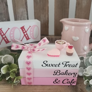 May include: A stack of decorative books in white and pink, tied with a pink ribbon featuring heart patterns. The books read "Sweet Treat Bakery & Cafe." A pink creamer, coffee cup, cupcake, and heart-shaped candy are on top. A sign with "XOXO" is in the background.