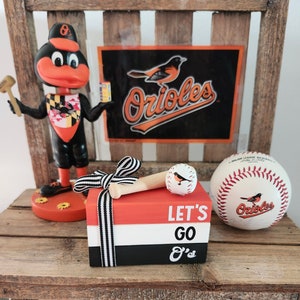 May include: A wooden box with black and white stripes and the words "Let's Go O's" on it. The box is decorated with a baseball bat and a baseball with an orange bird on it. There is also a baseball with the words "Orioles" on it and an orange bird figurine with a Maryland flag on its chest.