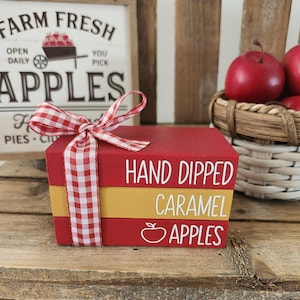 May include: A stack of three red and gold wooden blocks tied with a red and white checkered ribbon. The blocks read "HAND DIPPED CARAMEL APPLES" in white lettering. A basket of red apples is in the background.