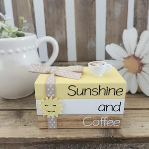 May include: A decorative stack of three books with the words "Sunshine and Coffee" printed on them. A small white coffee cup sits on top. A polka-dot ribbon and a smiling sun embellish the books. The books are yellow, white, and wood-toned.