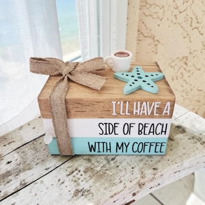 May include: A stack of three wooden blocks painted to resemble books, tied with a burlap ribbon. The blocks feature the text I'LL HAVE A SIDE OF BEACH WITH MY COFFEE. A small white coffee mug and a light blue starfish are placed on the top block.