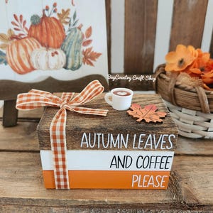 May include: A decorative wooden block set with the words "AUTUMN LEAVES AND COFFEE PLEASE" in white lettering. The set is adorned with a gingham ribbon, a miniature coffee cup, and a fall leaf. A pumpkin-themed painting is in the background.