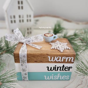 May include: A decorative wooden block set with the words "warm winter wishes" in white and black. The top block is brown, the middle is white, and the bottom is teal. A small blue coffee cup and a white snowflake are on top, along with a snowflake ribbon.