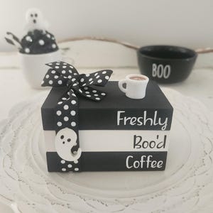 May include: A Halloween-themed decorative stack of books with the words "Freshly Boo'd Coffee" in white. A small white coffee cup sits on top. A black and white polka dot bow and ghost embellishments add to the spooky decor.