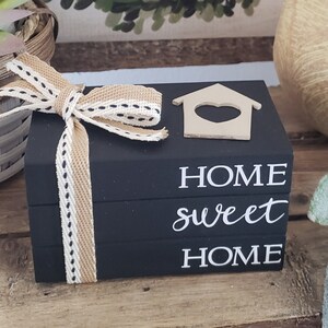 May include: Black decorative wooden blocks stacked with the words "HOME sweet HOME" in white script. A small wooden house with a heart cutout sits on top. A beige and white ribbon with black stitching is tied around the blocks.