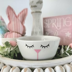 May include: A white ceramic bowl with a bunny face design, featuring closed eyelashes and a pink nose. The bowl is on a decorative tray with other Easter-themed items, including a pink bunny and a sign that says "SPRING".