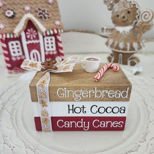 May include: A festive holiday display featuring a gingerbread house, a gingerbread person figurine, and a stack of books with the words "Gingerbread", "Hot Cocoa", and "Candy Canes". A small cup of hot cocoa and a candy cane are also present.