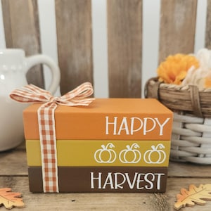 May include: A stack of three rectangular blocks resembling books, colored orange, yellow, and brown, tied with a brown and white gingham ribbon. The top block features the white text HAPPY, the middle block has three white pumpkin icons, and the bottom block features the white text HARVEST.